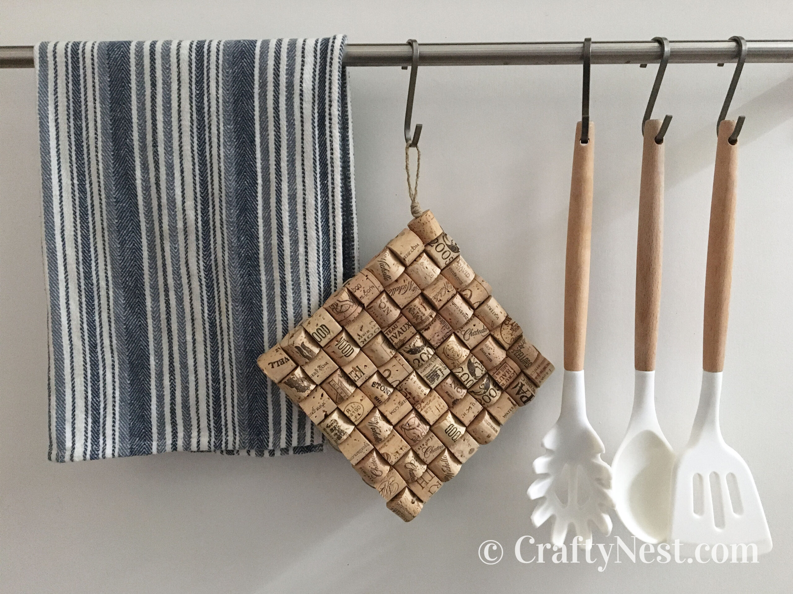 hanging-woven-wine-cork-trivet-crafty-nest Wine-cork trivet hanging from a hook with a dish towel and cooking utensils, photo